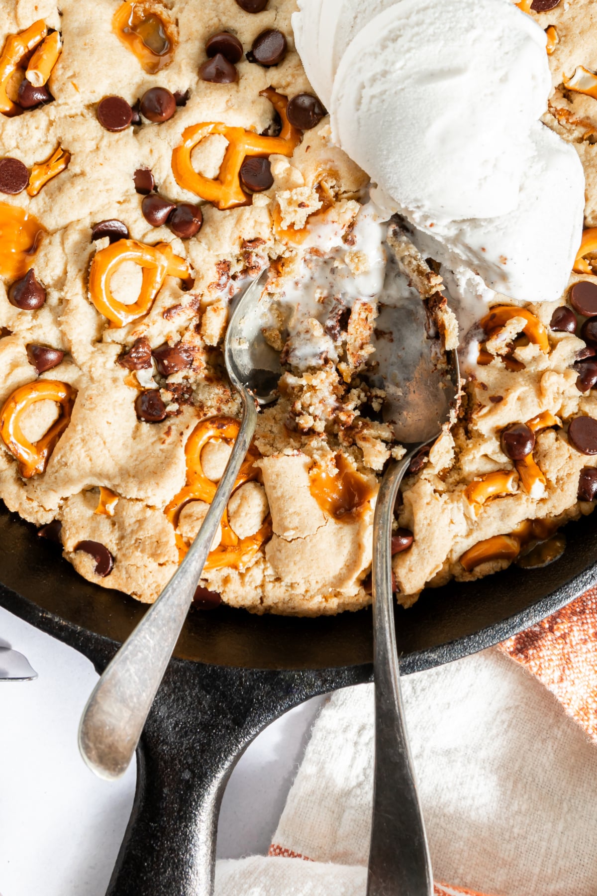 Overhead view of a baked skillet cookie with caramel, pretzels, and chocolate chips. Three scoops of vanilla ice cream melting on top, with two spoons digging into the cookie.