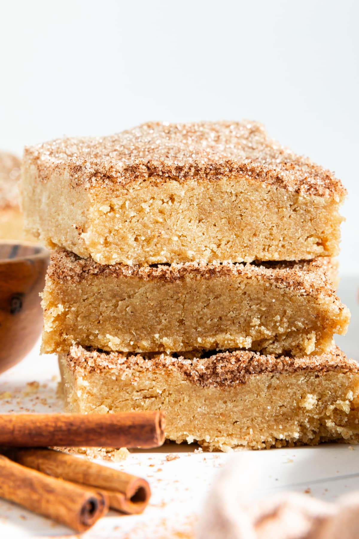 Three squares of snickerdoodle blondies stacked on top of each other. Blondies are topped with a cinnamon sugar mixture, and sit next to cinnamon sticks.