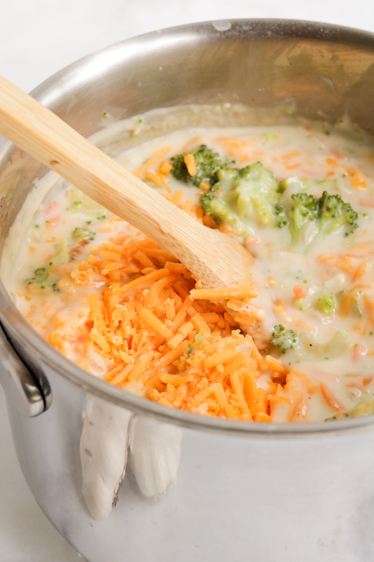 Close up of stainless steel pot of soup being made; cheese and broccoli are being stirred in with a wooden spoon.