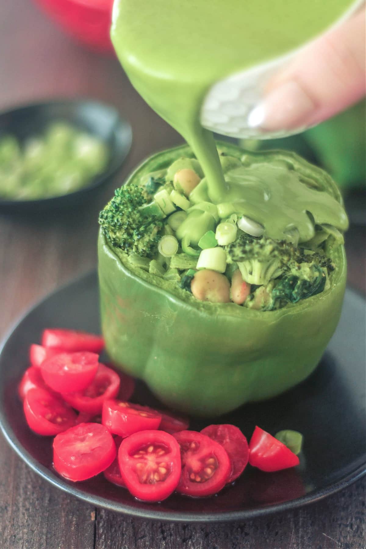 A hand pouring green sauce over a green bell pepper standing on a matte black plate. The pepper is stuffed with a green goddess mixture of beans, broccoli, onion, riced palm hearts. Sliced red cherry tomatoes sit on side of pepper as a salad / garnish.