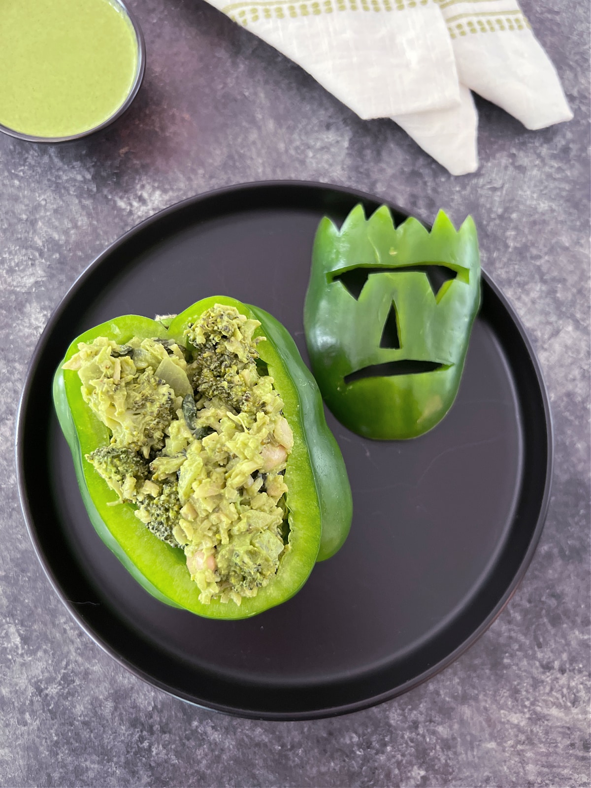 Overhead view of a green bell pepper that has been carved with a Frankenstein monster face and is stuffed with a green goddess sauce and vegetable mixture. The monster face is removed and sits on the black plate next to the pepper, which shows the green veggie stuffing inside.