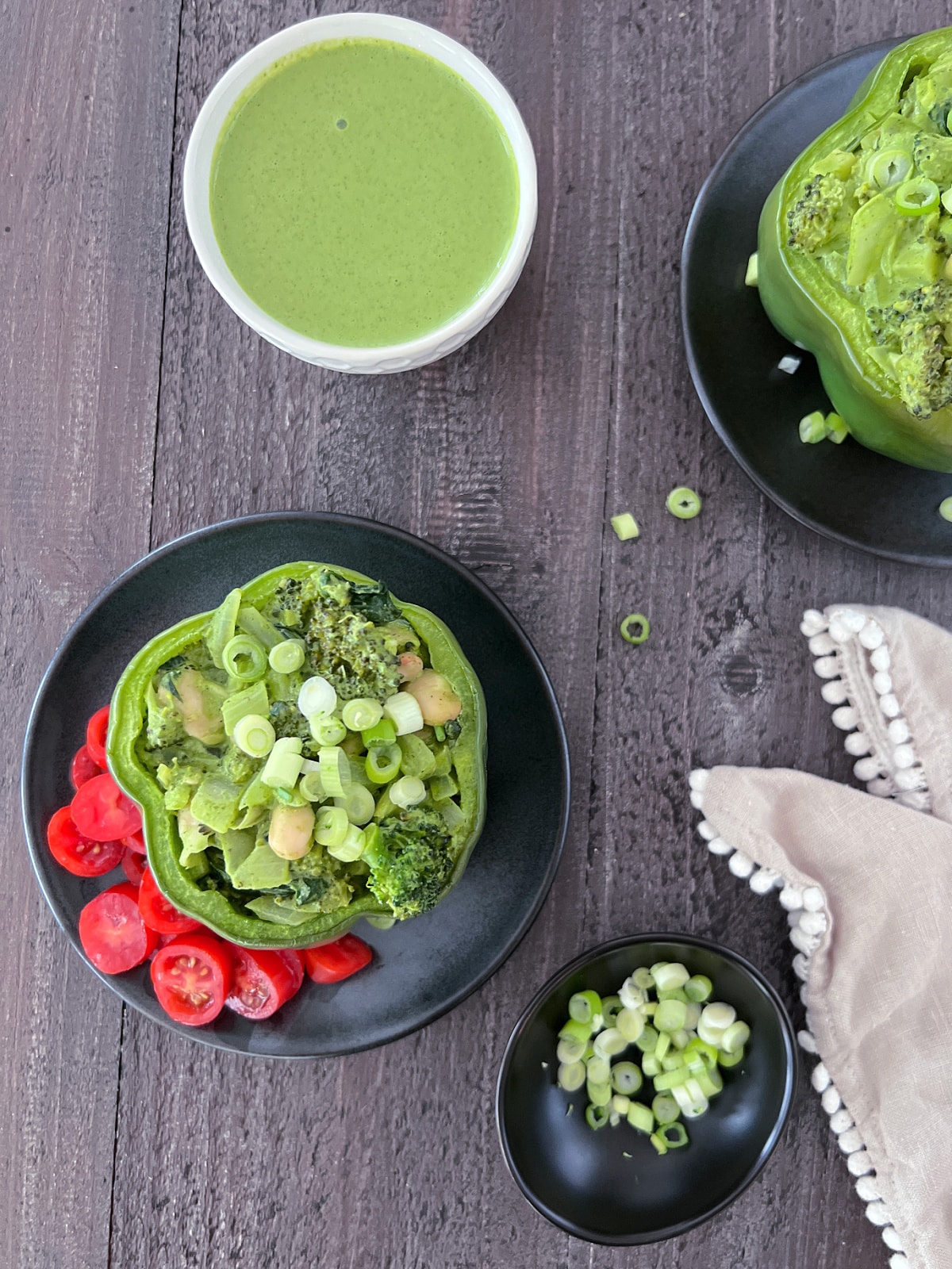 Overhead view of two green bell peppers standing on matte black plates, tops sliced off and stuffed with a green goddess mixture of beans, broccoli, onion, riced palm hearts, and green goddess sauce. One plate is garnished with sliced red cherry tomatoes.