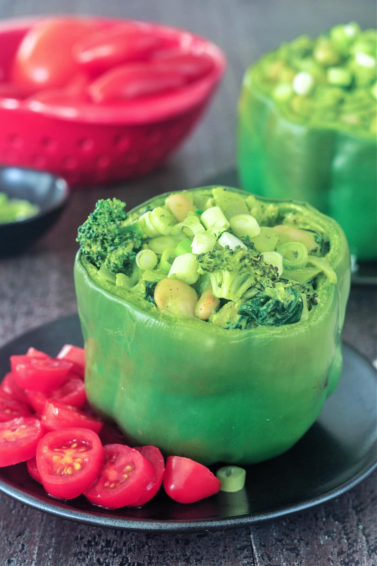 Two green bell peppers standing on matte black plates, tops sliced off and stuffed with a green goddess mixture of beans, broccoli, onion, riced palm hearts, and green goddess sauce. One plate is garnished with sliced red cherry tomatoes.