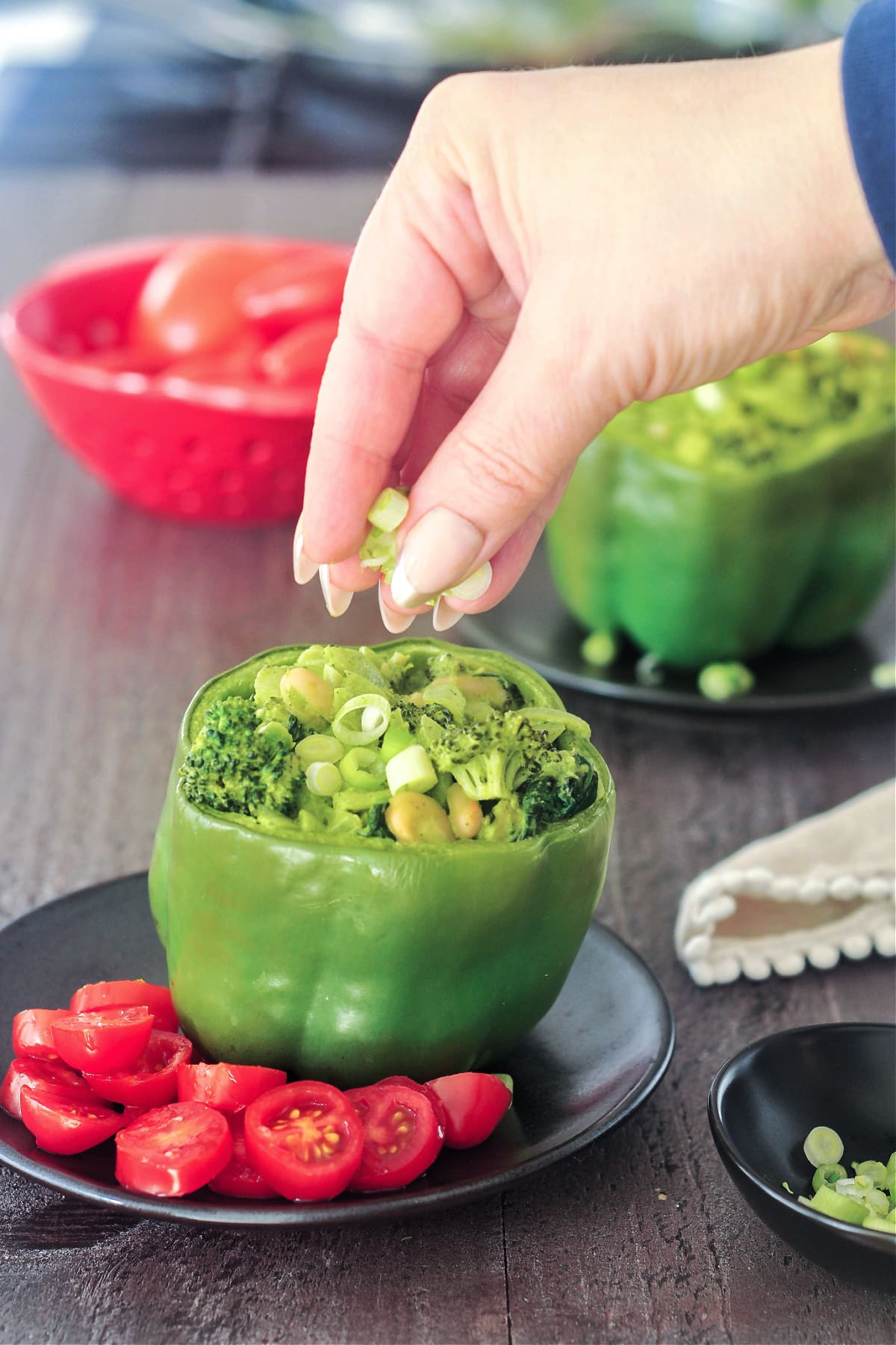 A hand sprinkling sliced green onions over a green bell pepper standing on a matte black plate. The pepper is stuffed with a green goddess mixture of beans, broccoli, onion, riced palm hearts. Sliced red cherry tomatoes sit on side of pepper as a salad / garnish.