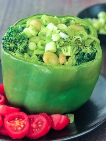 A green bell pepper standing on a matte black plate, tops sliced off and stuffed with a green goddess mixture of beans, broccoli, onion, riced palm hearts, and green goddess sauce. Sliced red cherry tomatoes sit on side of pepper as a salad / garnish.