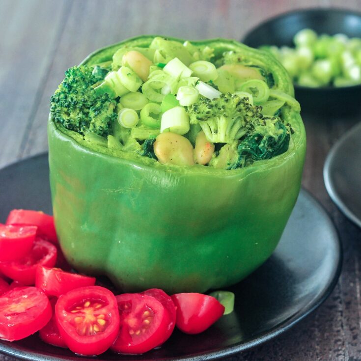 A green bell pepper standing on a matte black plate, tops sliced off and stuffed with a green goddess mixture of beans, broccoli, onion, riced palm hearts, and green goddess sauce. Sliced red cherry tomatoes sit on side of pepper as a salad / garnish.