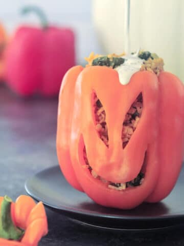 An orange bell pepper carved with a Halloween jack o lantern face, then filled with vegan buffalo chicken, rice, and veggies. A hand is pouring ranch dressing onto the top of the bell pepper.