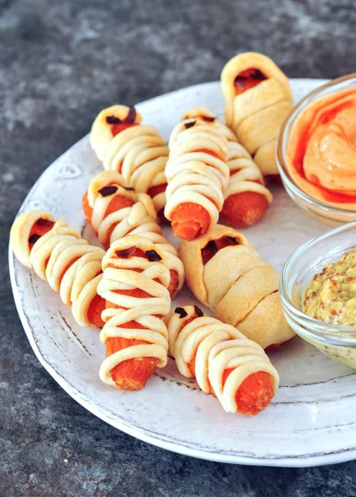 Mini carrots wrapped in pastry to look like mummies for Halloween sit on a serving plate with small glass bowls of hot sauce and mustard.