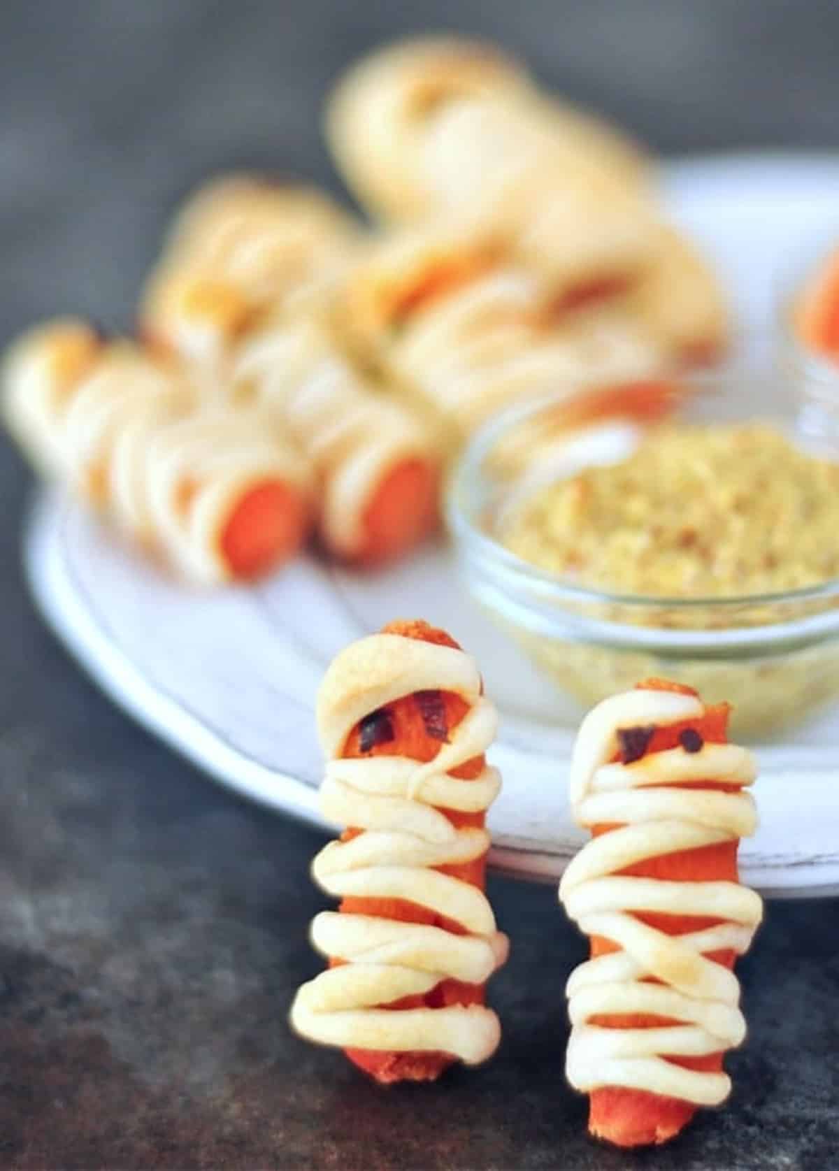 Mini carrots wrapped in pastry to look like mummies for Halloween sit on a serving plate with small glass bowls of hot sauce and mustard. Two "mummies" stand at the edge of the plate.