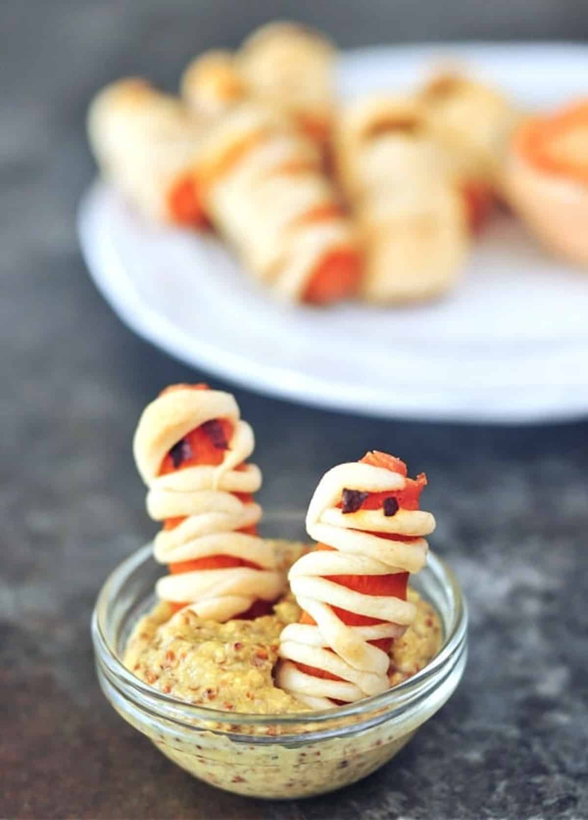 Mini carrots wrapped in pastry to look like mummies for Halloween sit on a serving plate with small glass bowls of hot sauce and mustard. Two "mummies" stand inside the bowl of mustard.