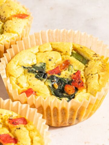 Close up of mini vegan quiche on an off white countertop. The vegan egg bites are bright yellow from turmeric, and are filled with red peppers, spinach, and asparagus.