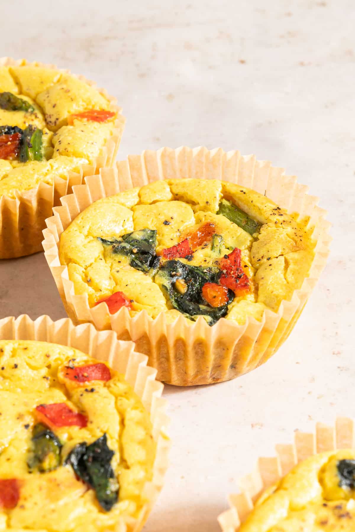 Four mini vegan egg cups baked in cupcake liners sit on an off white surface. The vegan egg bites are bright yellow from turmeric, and are filled with red peppers, spinach, and asparagus.