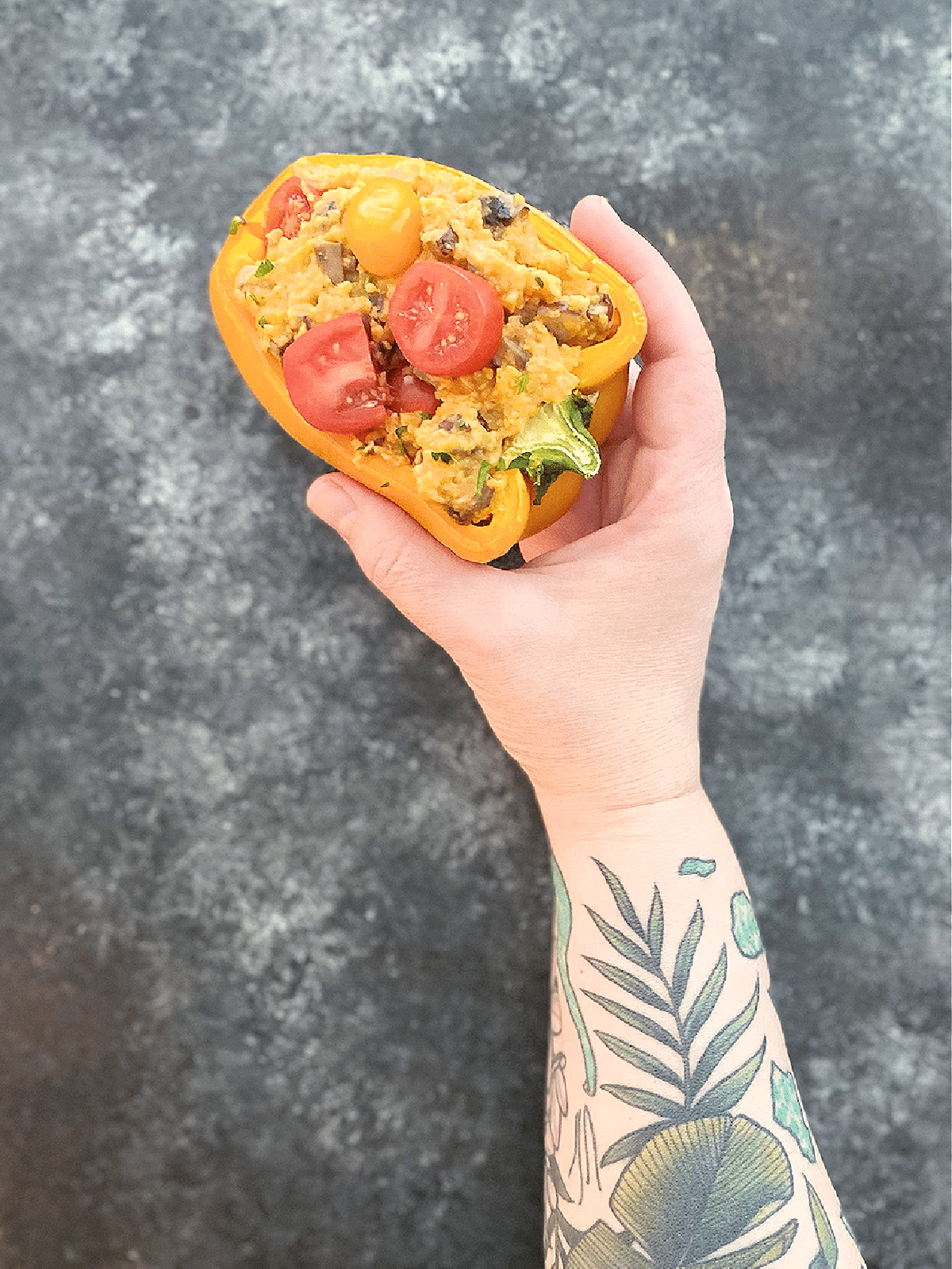 A hand holding a half orange bell pepper stuffed with pizza flavored mushroom polenta, topped with sliced cherry tomatoes. Arm is tattooed with tropical leaves and the Hawaiian islands.