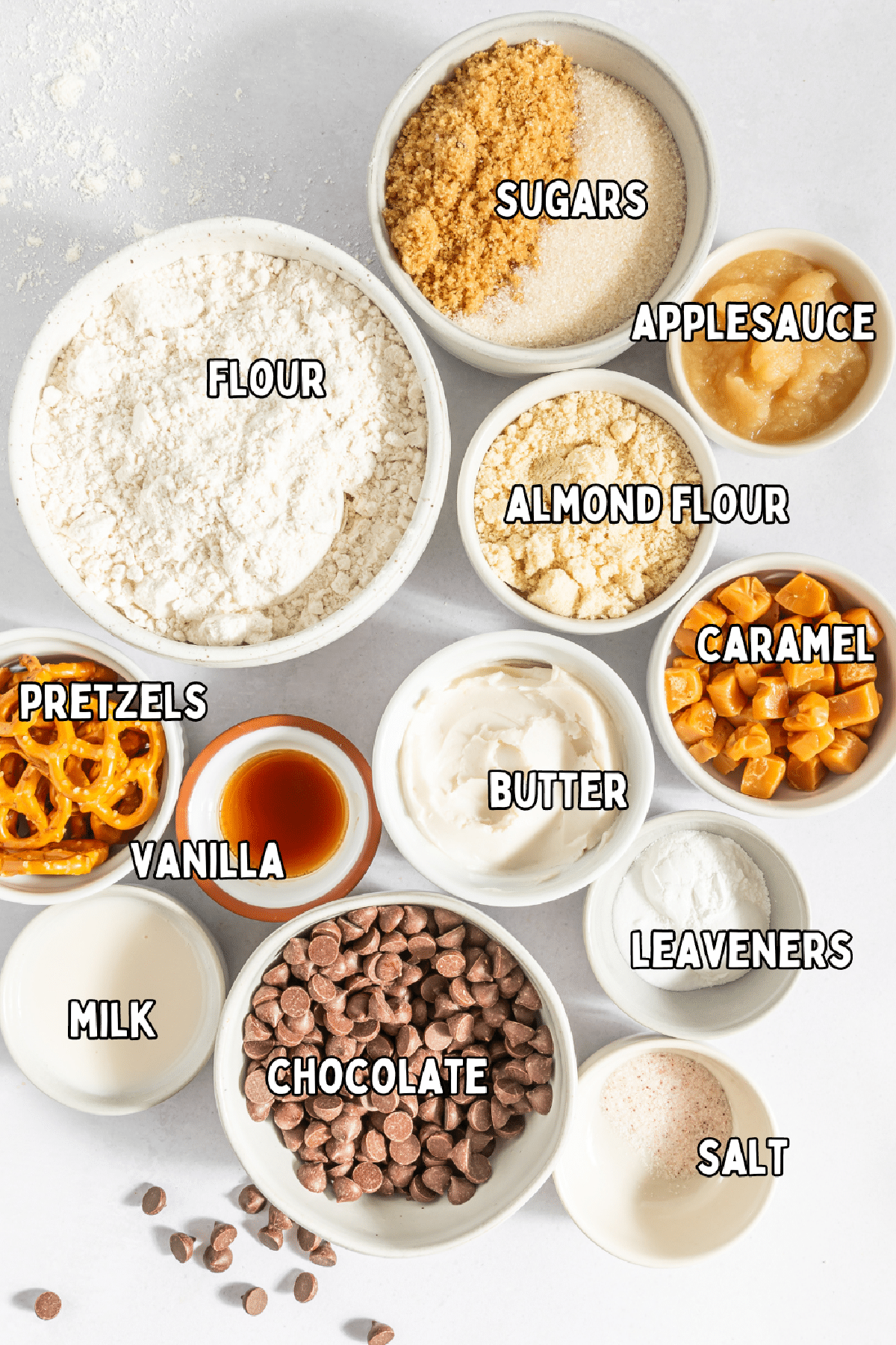Small bowls of ingredients for a skillet cookie: flours, sugars, applesauce, caramel, butter, vanilla, pretzels, milk, chocolate, leaveners, and salt.