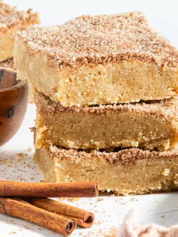 Close up of three squares of snickerdoodle blondies stacked on top of each other. Blondies are topped with a cinnamon sugar mixture, and sit next to a small wooden bowl of cinnamon sugar and cinnamon sticks.