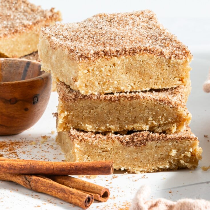 Close up of three squares of snickerdoodle blondies stacked on top of each other. Blondies are topped with a cinnamon sugar mixture, and sit next to a small wooden bowl of cinnamon sugar and cinnamon sticks.