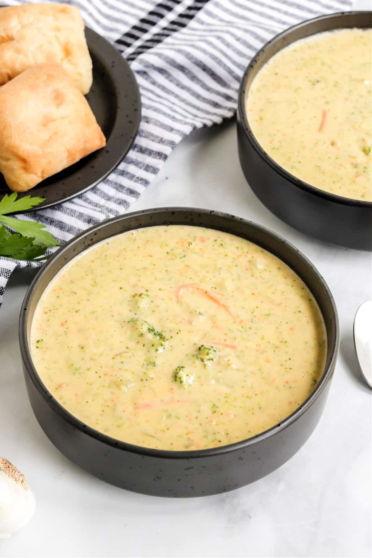 Broccoli cheese soup in two matte black bowls. A black plate of golden toasted rolls sit next to the soup on a grey and white striped cloth napkin.