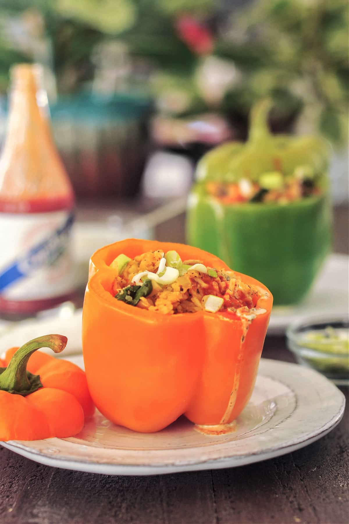 An orange bell pepper filled with vegan buffalo chicken and rice mixture. A green stuffed bell pepper is blurred in background, along with a bottle of hot sauce.