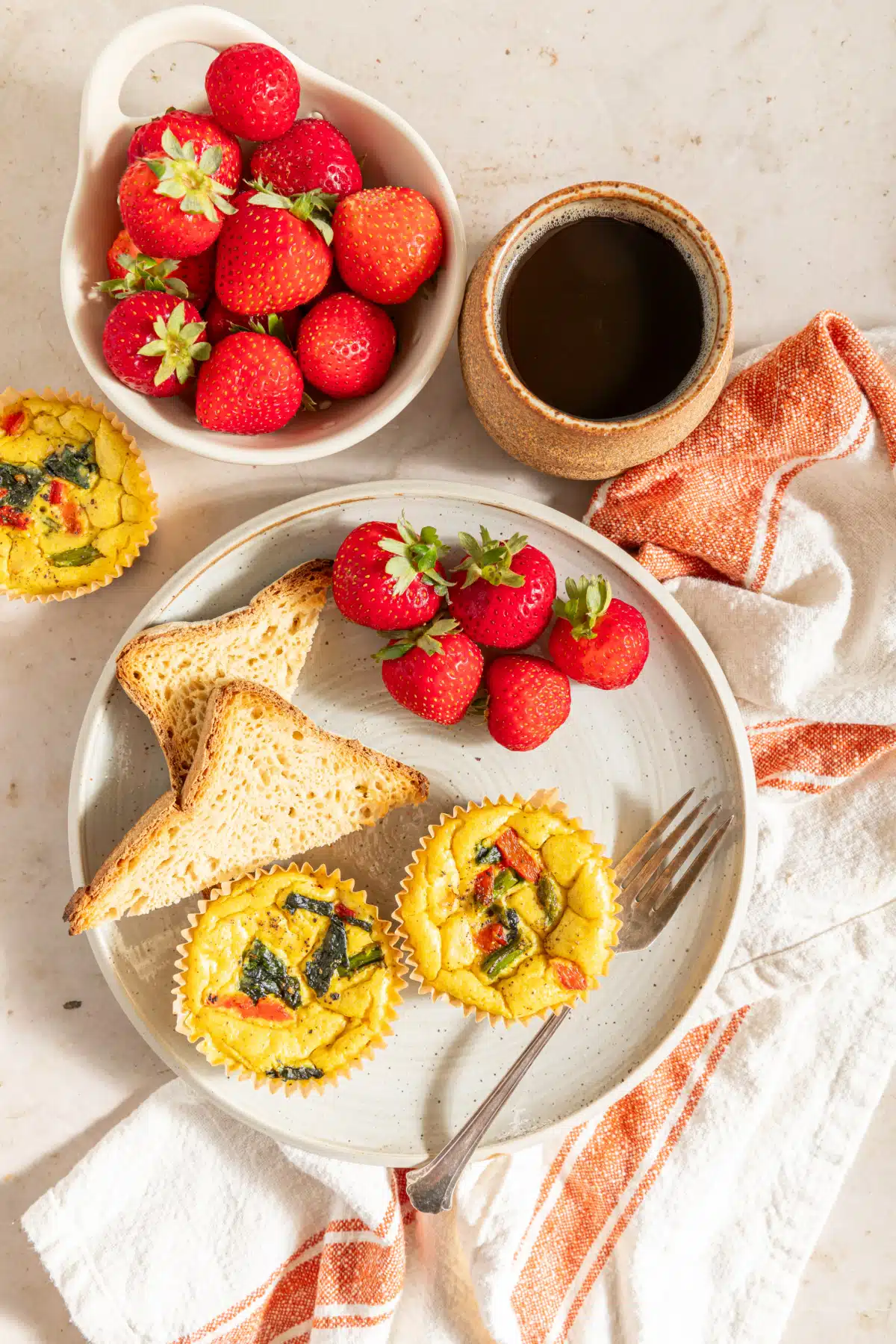 Overhead view of two mini vegan quiche baked in cupcake liners on a white plate with a fork, strawberries, and toast. The vegan egg bites are bright yellow from turmeric, and have red peppers, spinach, and asparagus in them. A bowl of strawberries and a cup of coffee sit next to the plate with a white and orange cloth napkin.