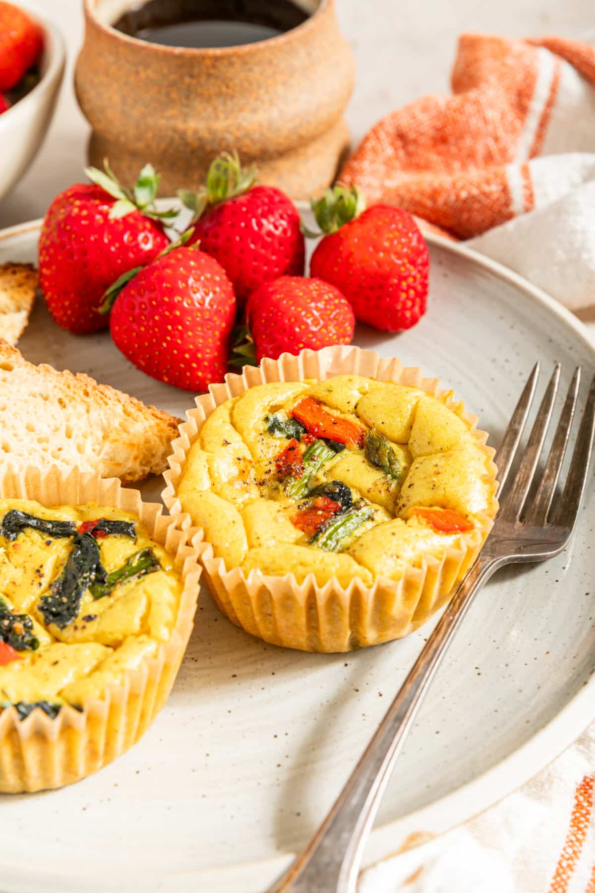 Two mini vegan quiche baked in cupcake liners are on a white plate with a fork, strawberries, and toast. The vegan egg bites are bright yellow from turmeric, and have red peppers, spinach, and asparagus in them.