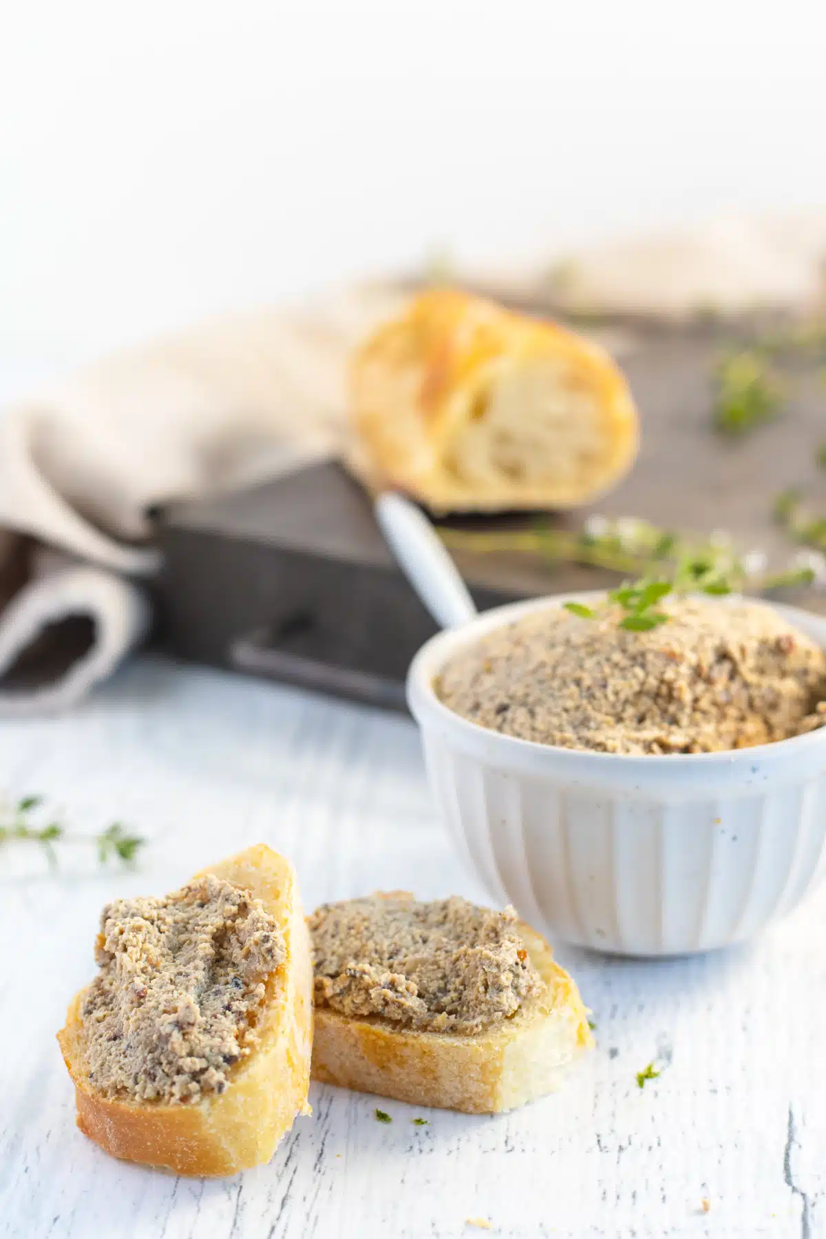 Vegan walnut pate in a white ceramic bowl, with sliced baguette bread on a cutting board blurred in background. Two slices of bread with pate sit in front of the pate bowl.