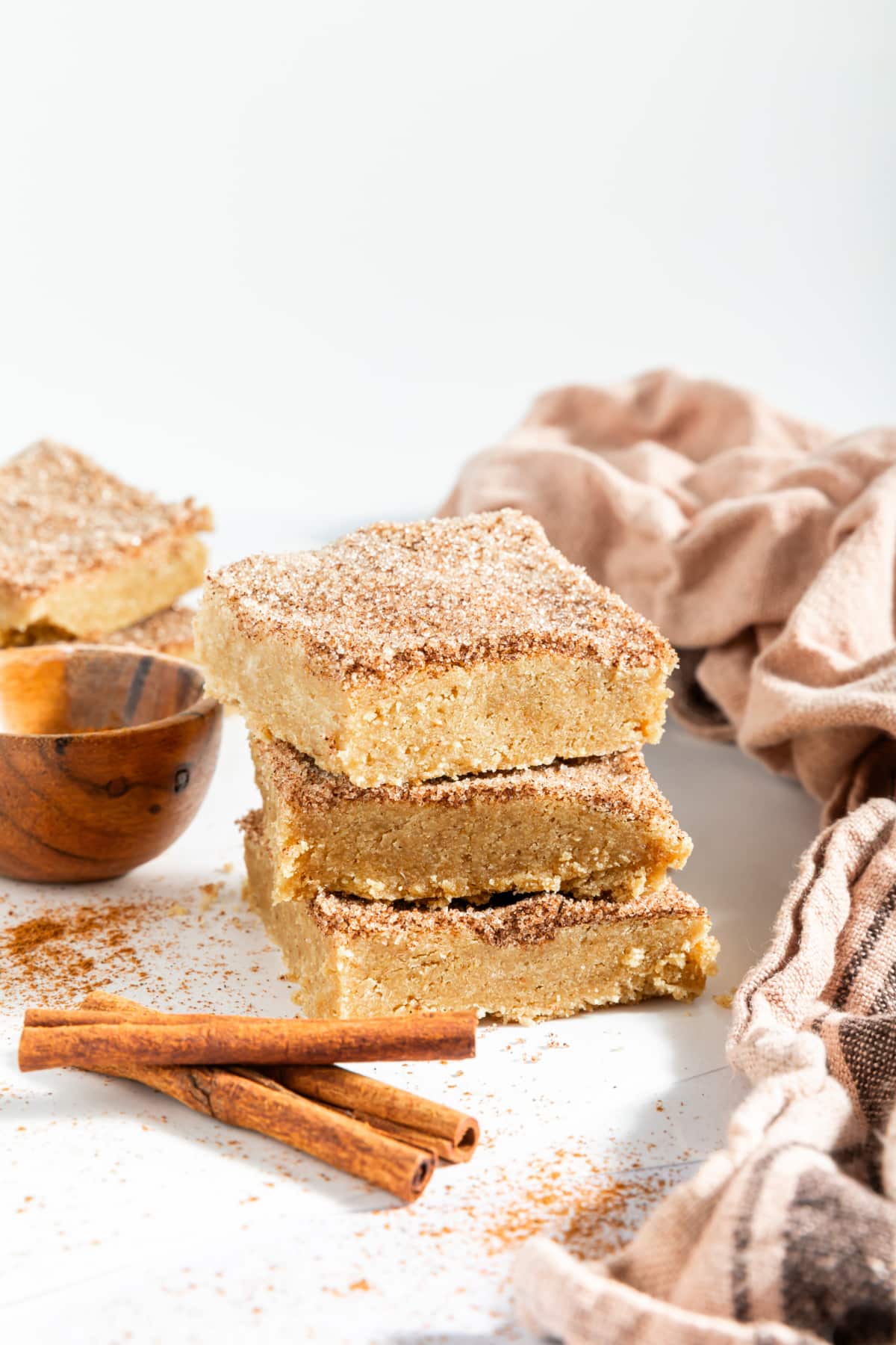 Three squares of snickerdoodle blondies stacked on top of each other. Blondies are topped with a cinnamon sugar mixture, and sit next to a small wooden bowl of cinnamon sugar and cinnamon sticks. A beige cloth napkin is crumpled next to the cookie bar stack.