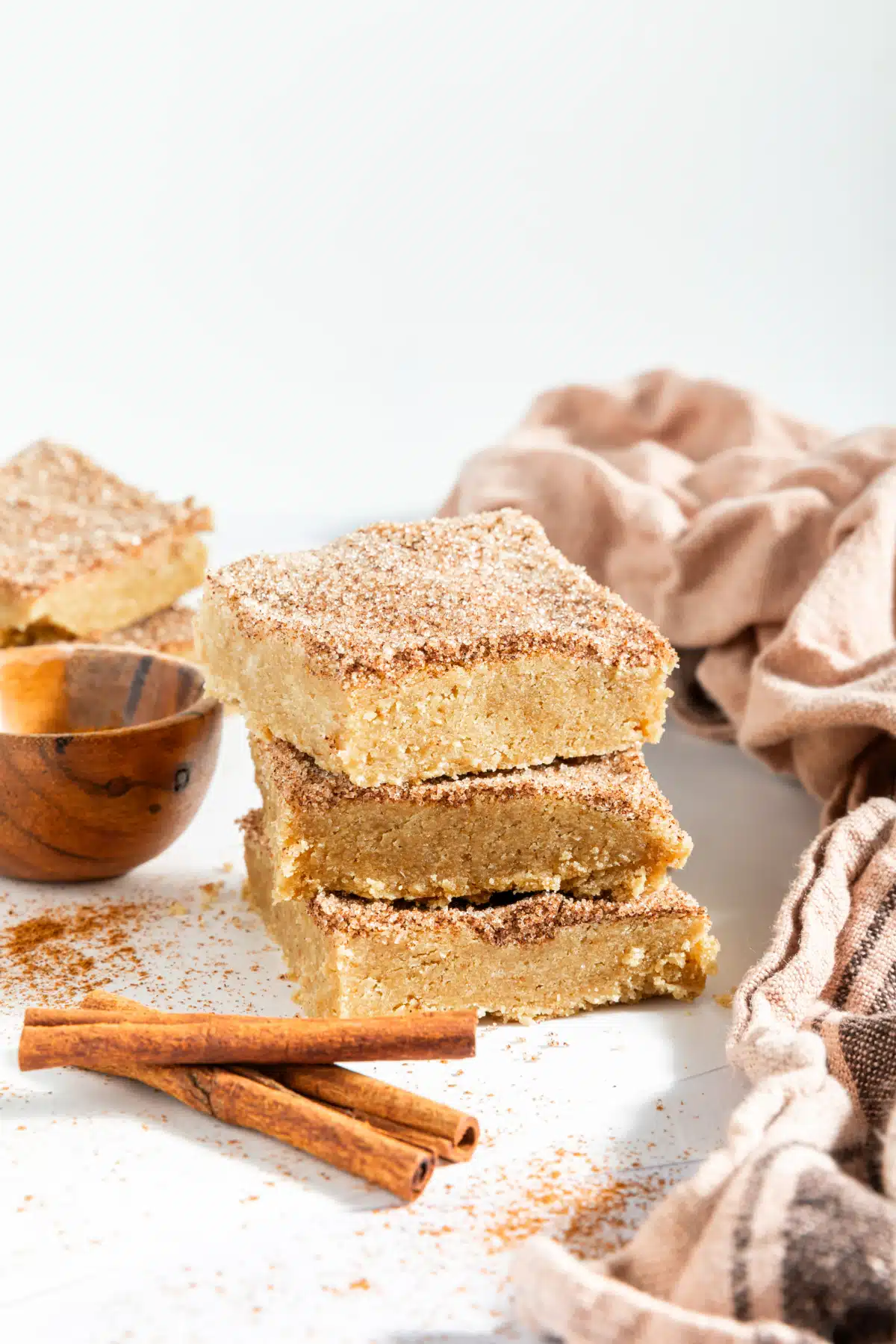 Three squares of snickerdoodle blondies stacked on top of each other. Blondies are topped with a cinnamon sugar mixture, and sit next to a small wooden bowl of cinnamon sugar and cinnamon sticks. A beige cloth napkin is crumpled next to the cookie bar stack.