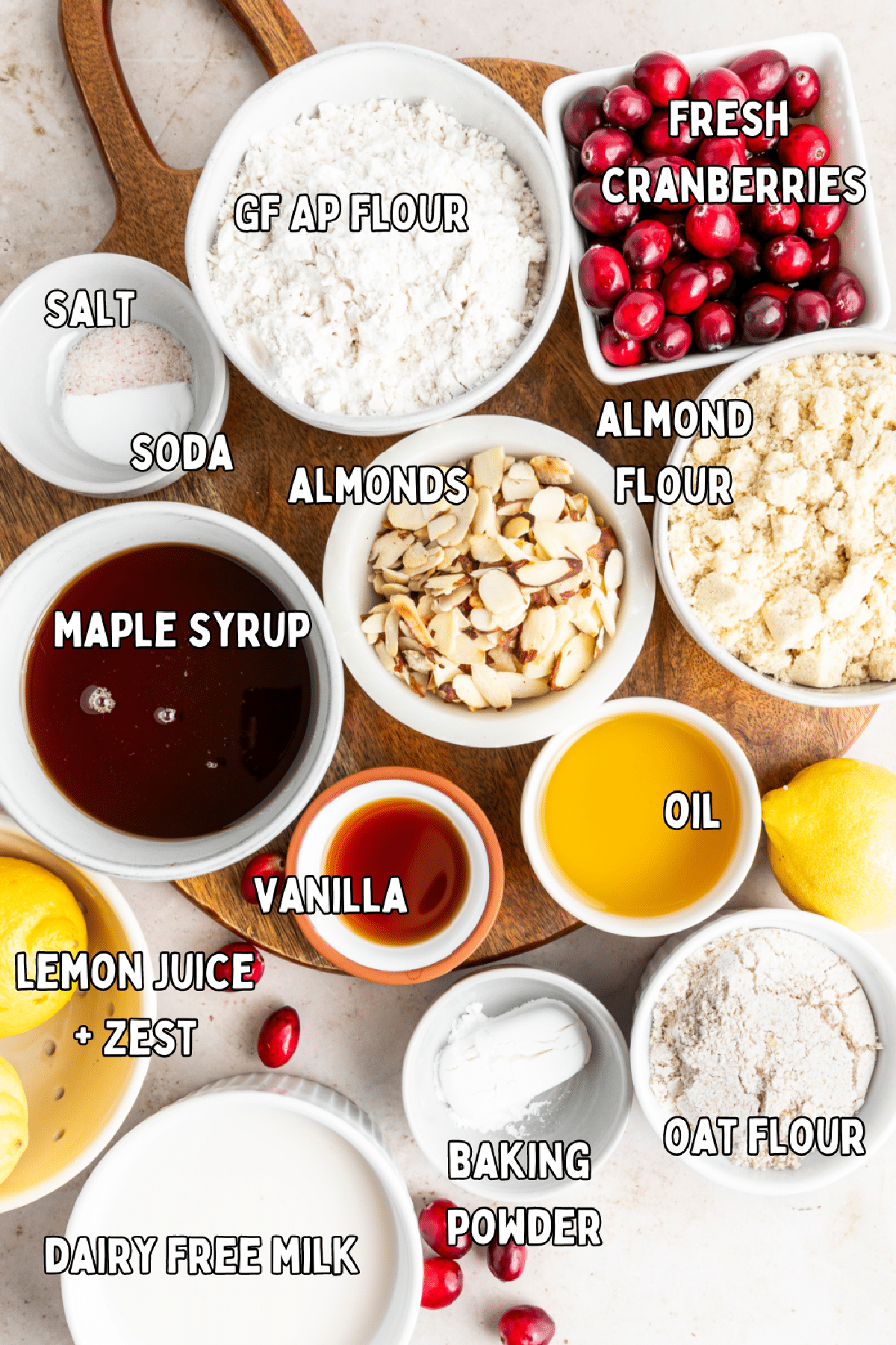 Overhead view of bowls with ingredients for cranberry lemon muffins: flours, fresh cranberries, sliced almonds, maple syrup, vanilla, oil, salt, lemon juice and zest, baking powder and soda.