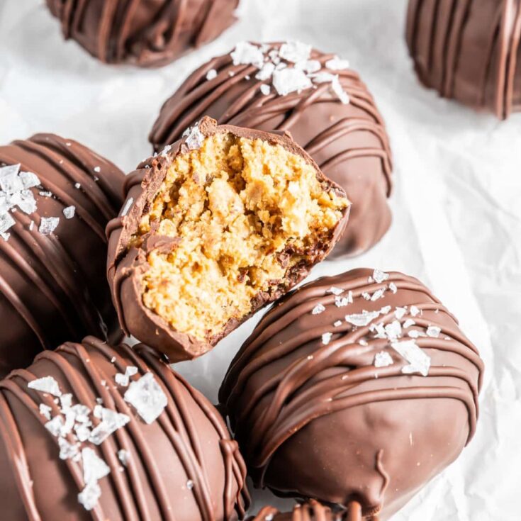 chocolate coated peanut butter crunch balls on white parchment paper. One ball is cut in half to show crispy insides.