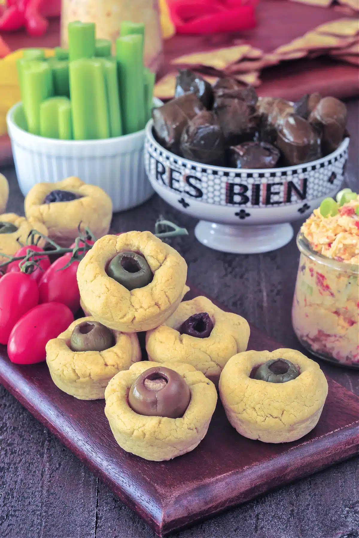 Cheesy olive bites on a dark wood serving board with cherry tomatoes. Olive board is sitting next to a small bowl of stuffed grape leaves, a jar of pimiento cheese spread, a bowl of celery sticks, and another snack board of vegetables.