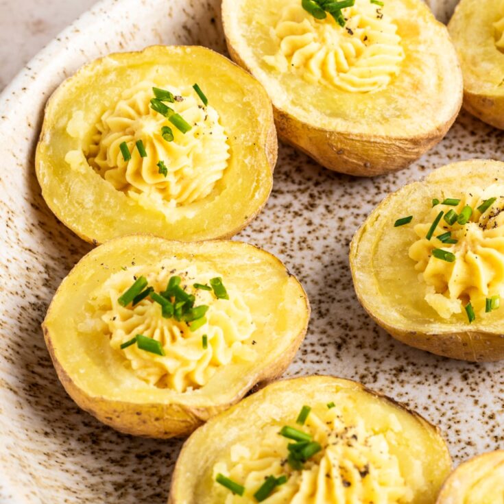 Close up view of mini gold potatoes sliced in half, baked, scooped out and filled with a deviled egg flavored mixture. These deviled potatoes are sitting on a tan speckled dish for serving.