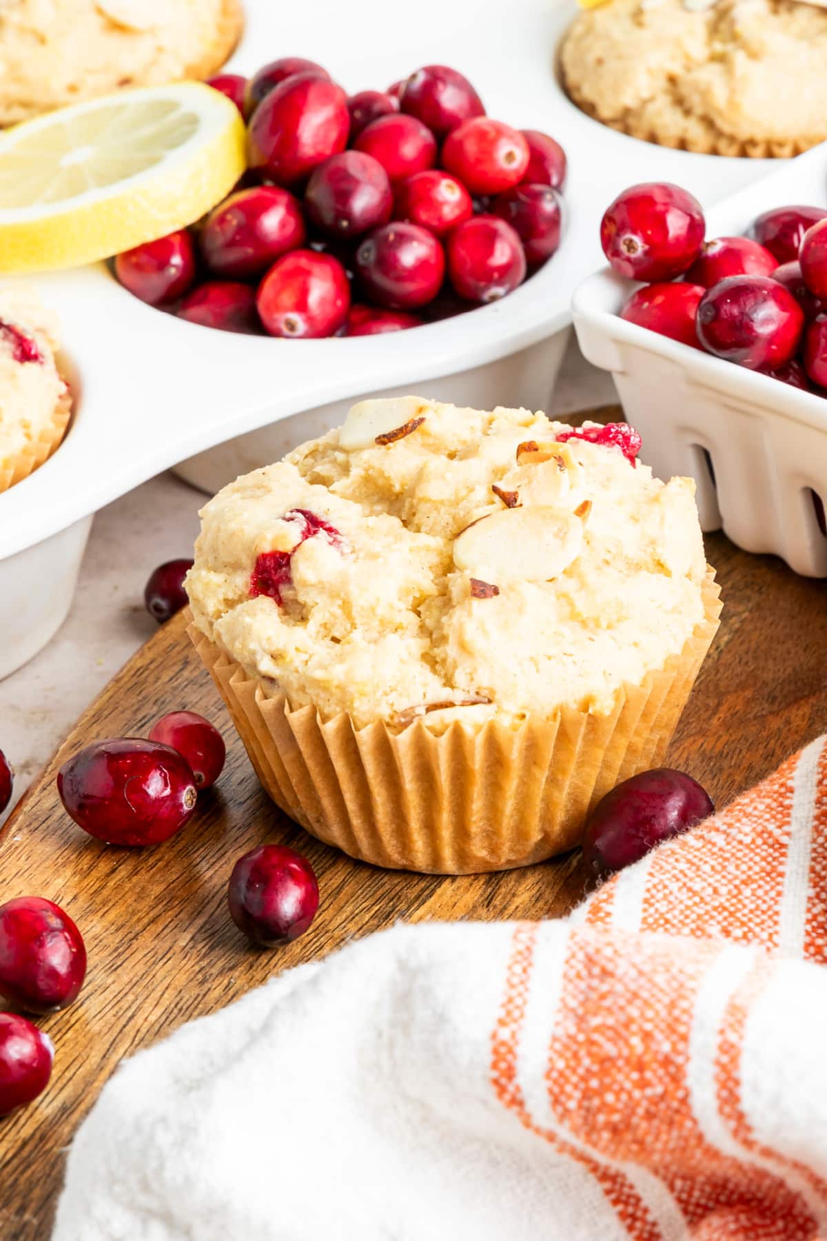 A gluten free cranberry muffin sitting on a wood board with a ceramic berry box full of fresh cranberries and an orange and white tea towel. A white muffin tin full of fresh baked muffins sits to the left.