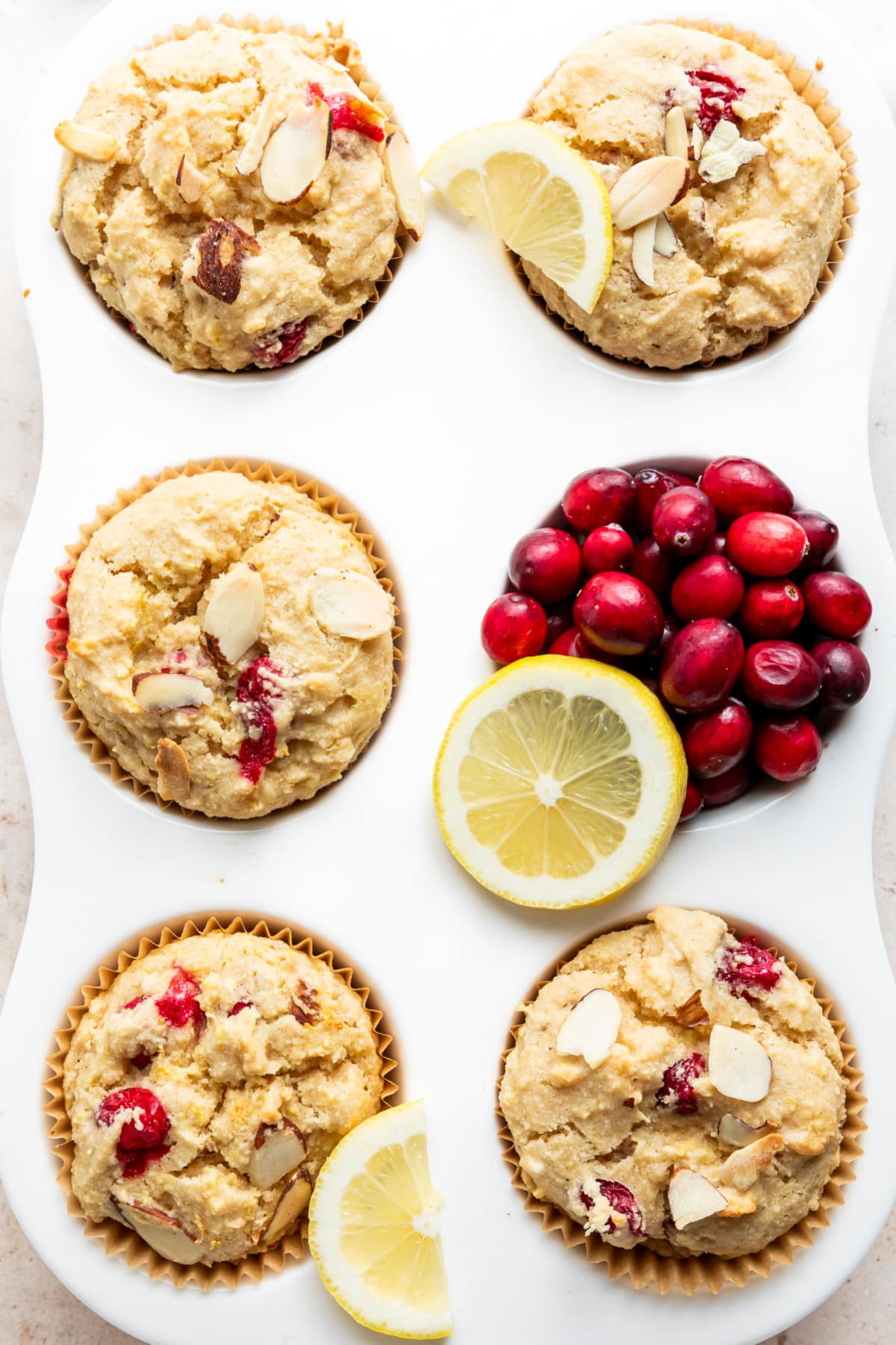 Overhead view of a 6 cup white muffin tin full of fresh baked muffins. There are five cranberry lemon muffins and one muffin cup full of fresh cranberries. Slices of lemon sit on two of the muffins and on the side of the cranberries.