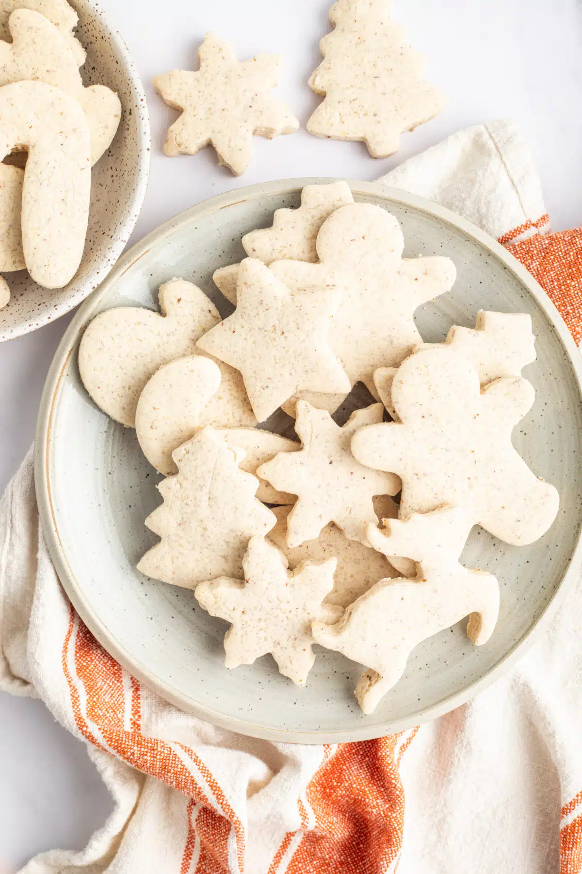 Overhead view of unfrosted, baked cut out sugar cookies arranged on plate. An orange and white striped kitchen towel is under the plate, and a couple cut out cookies are sitting off to the side.