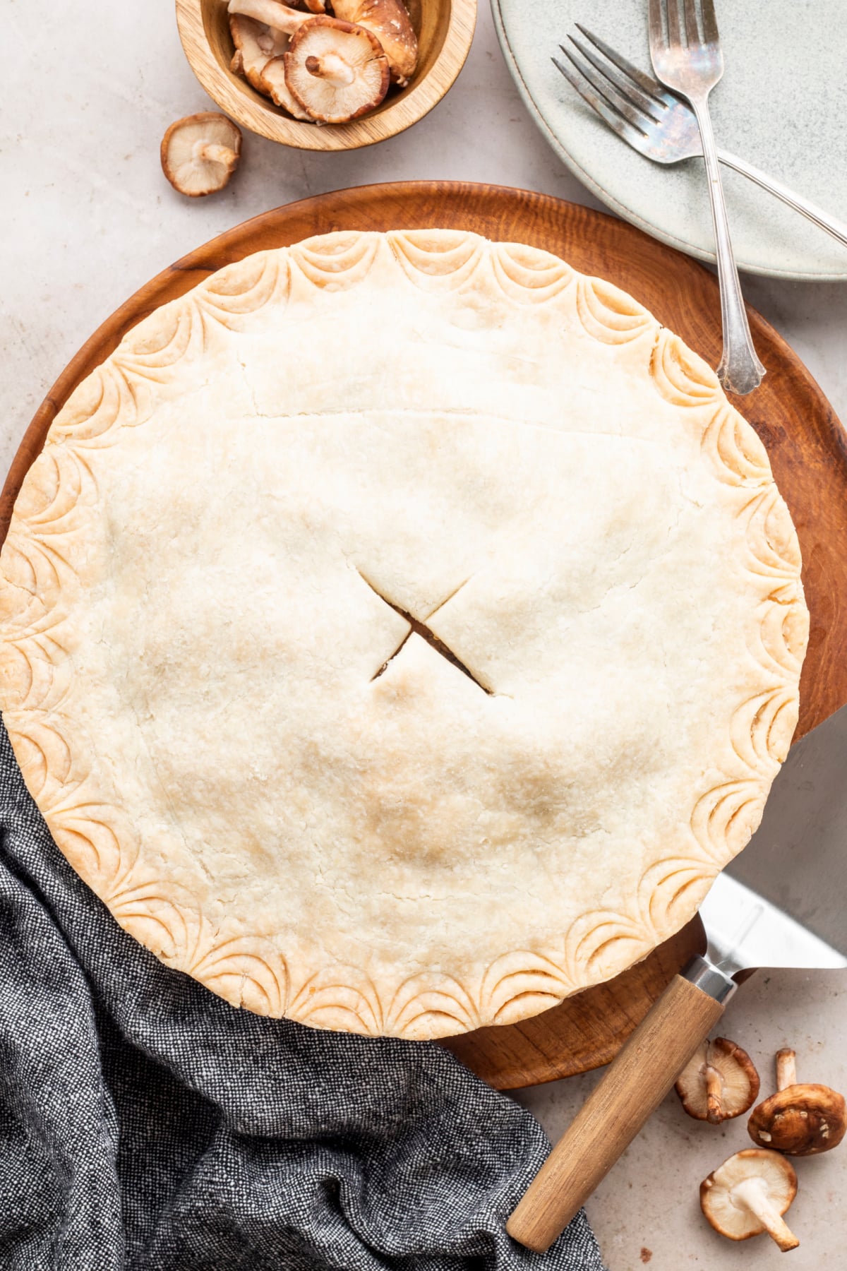 Overhead view of a whole baked pie with a golden tan top crust with a slice in the middle and decorated edging. A grew cloth napkin sits on the side, along with a pie cutter, a stack of plates with forks, and a small wooden bowl of mushrooms, indicating the filling of this savory pie.