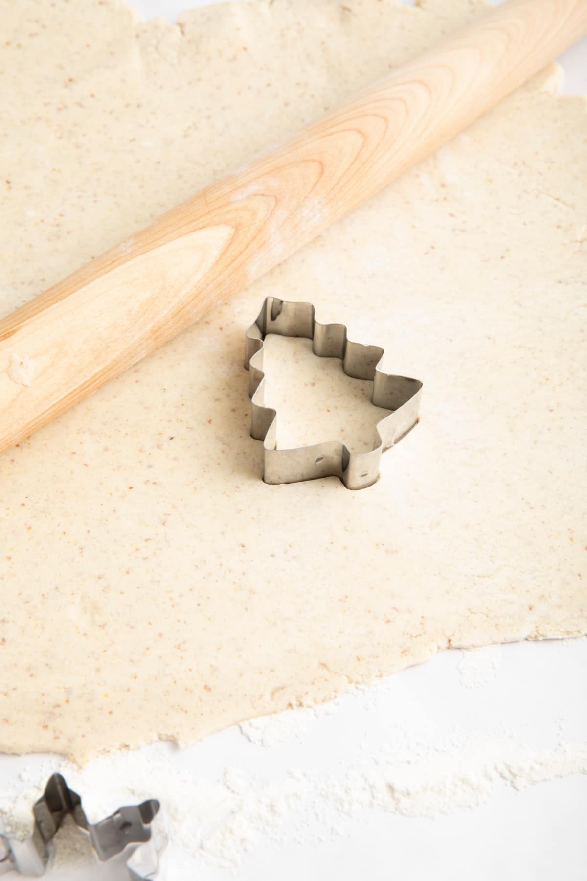 Sugar cookie dough rolled out on a floured white surface. A light wood rolling pin and a Christmas tree shaped cookie cutter sit on the dough. A snowflake cookie cutter sits just off to the side of the cookie dough.