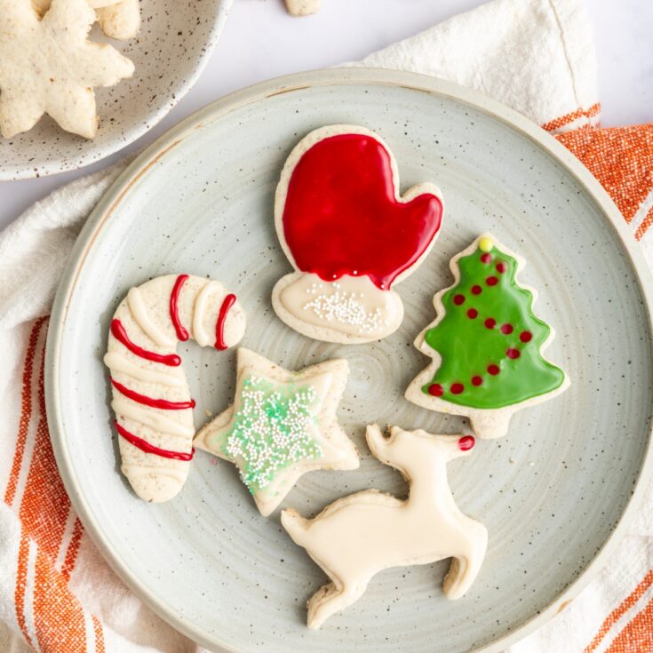 Frosted gluten free vegan cut out cookies in holiday shapes sit on a serving plate: a red mitten, a green Christmas tree with red dots, a white reindeer with a red nose, a red and white striped candy cane, and a green and white sprinkled star.