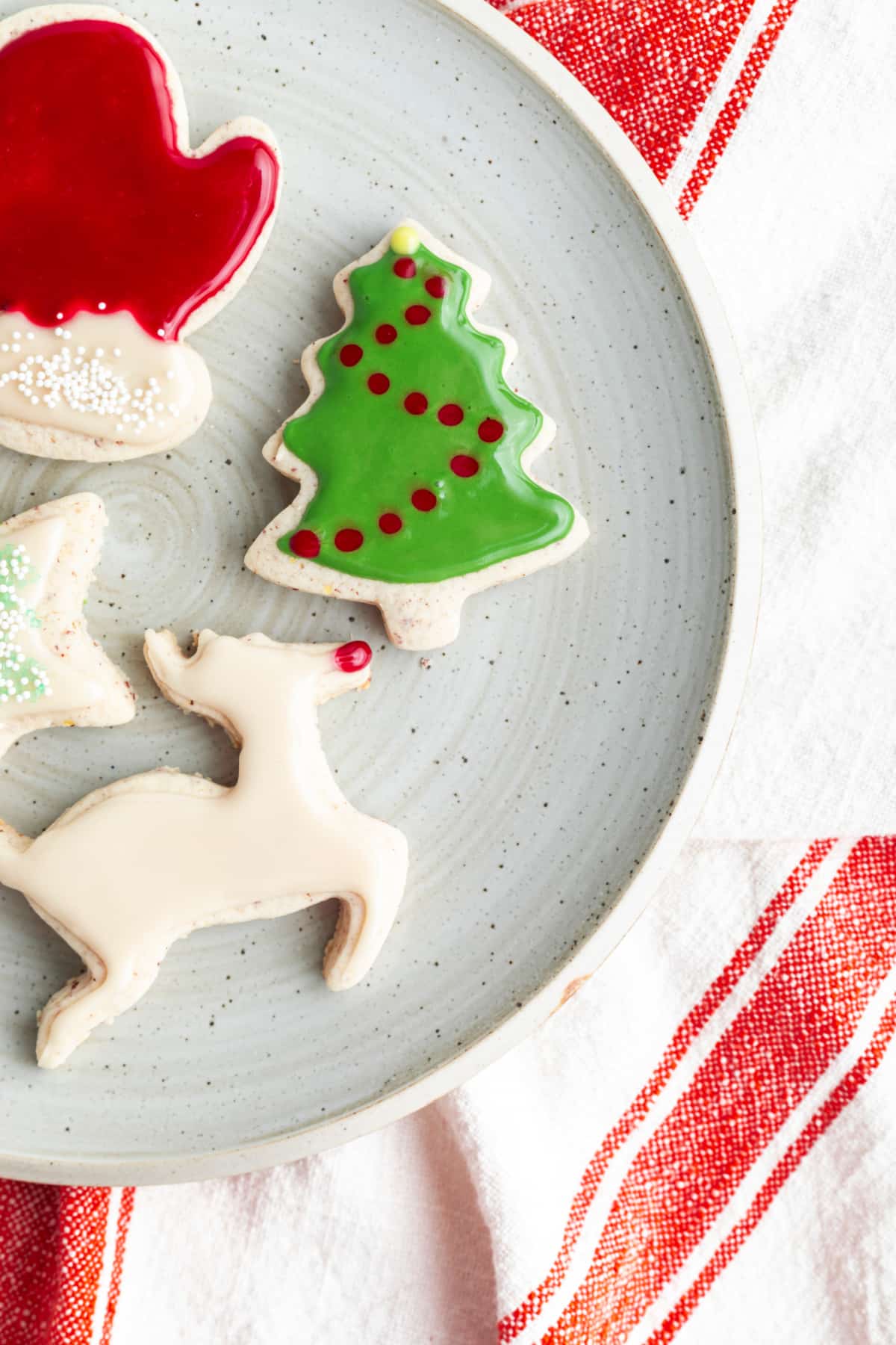 Frosted gluten free vegan cut out cookies in holiday shapes sit on a serving plate: a red mitten, a green Christmas tree with red dots, and a white reindeer with a red nose.