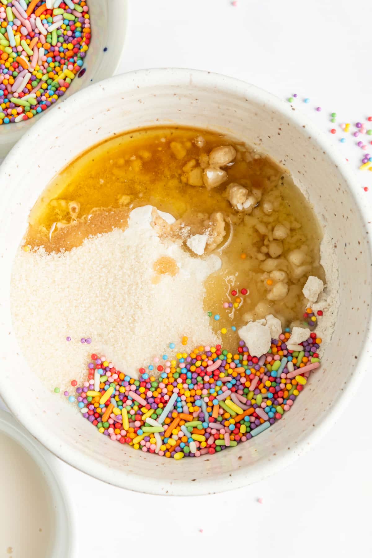 A white mixing bowl of muffin batter before mixing: melted butter, vanilla, flour, and sprinkles.