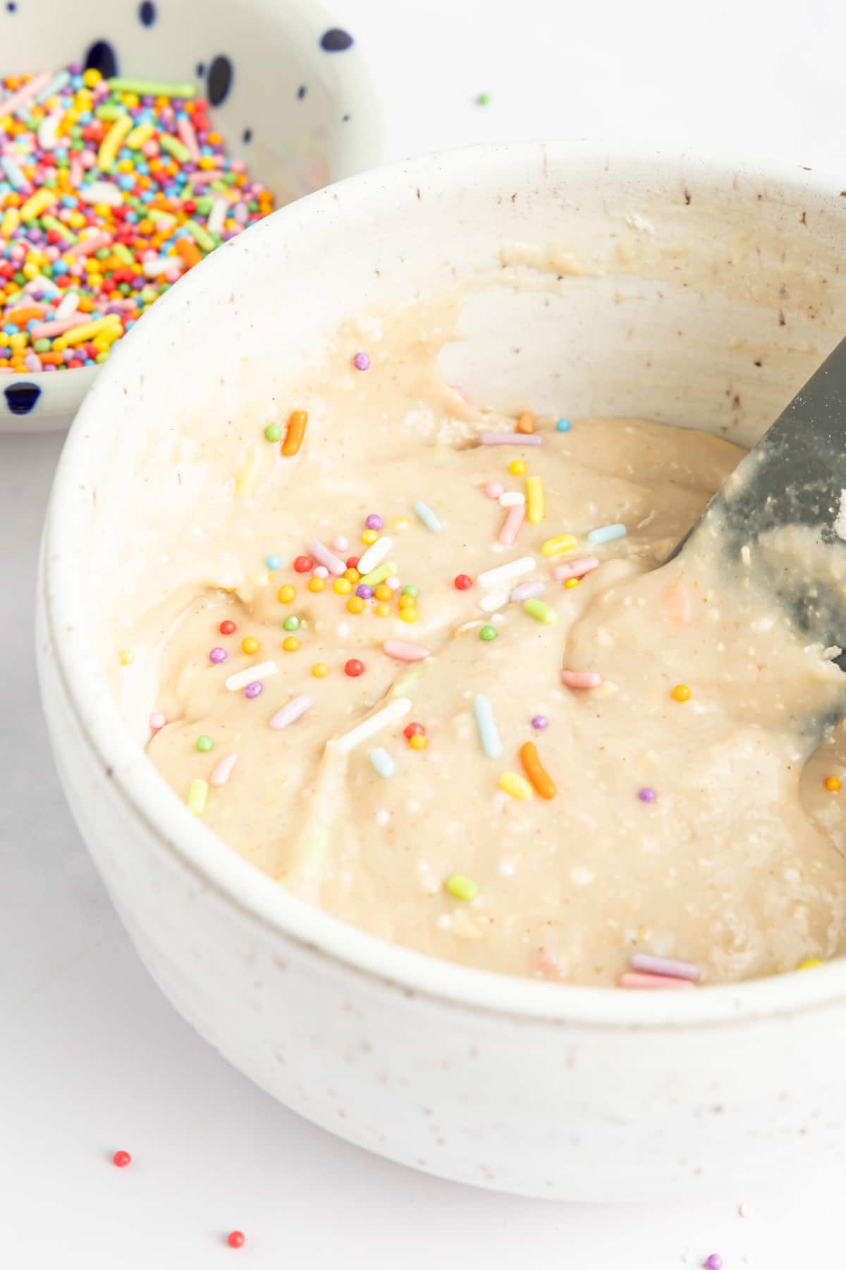 Pale tan muffin batter with pastel colored sprinkles in a white mixing bowl, sitting next to a small bowl of those pastel sprinkles.