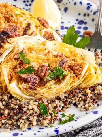 Maple mustard roasted cabbage steaks with breadcrumbs and pecans served over cooked quinoa and garnished with lemon wedges sit on a white and blue plate.