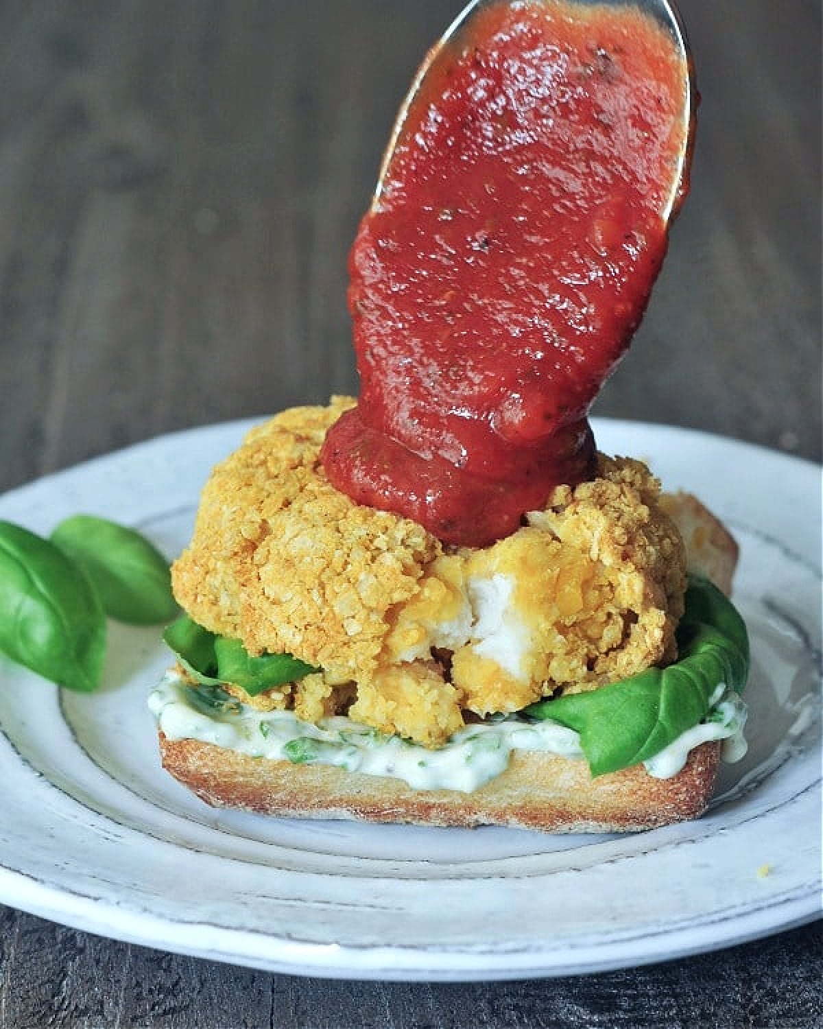 Marinara sauce is being spooned over a vegan chicken burger patty on a bottom bun with basil aioli and fresh basil leaves.