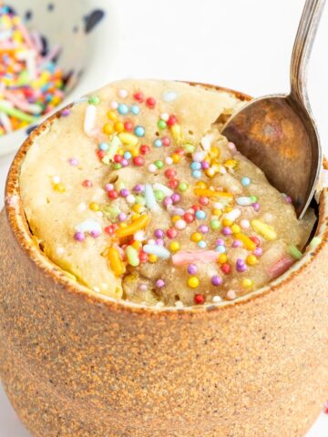 Close up of a vanilla mug cake in the light brown pottery mug it was cooked in. Cake is yellow with sprinkles and there is a spoon going into the cake.
