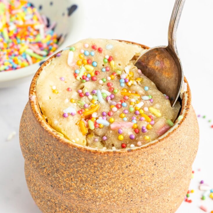 Close up of a vanilla mug cake in the light brown pottery mug it was cooked in. Cake is yellow with sprinkles and there is a spoon going into the cake.