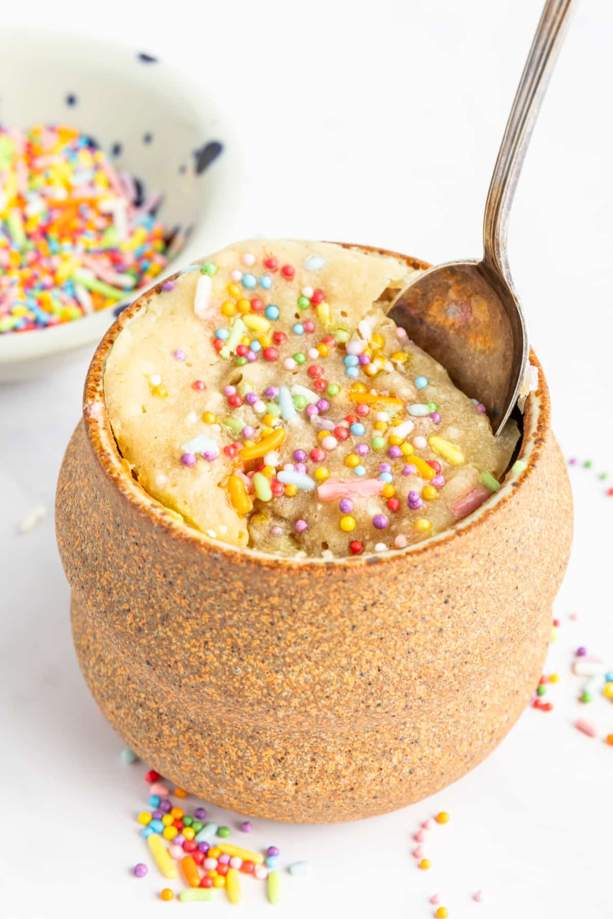 A spoon sits in a vanilla mug cake in the light brown pottery mug it was cooked in. Cake is yellow with sprinkles.