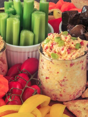 Pimiento cheese in two small glass jars, surrounded by veggies and crackers on a snack board.