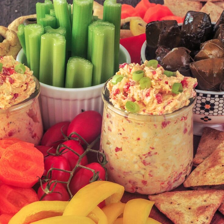 Pimiento cheese in two small glass jars, surrounded by veggies and crackers on a snack board.