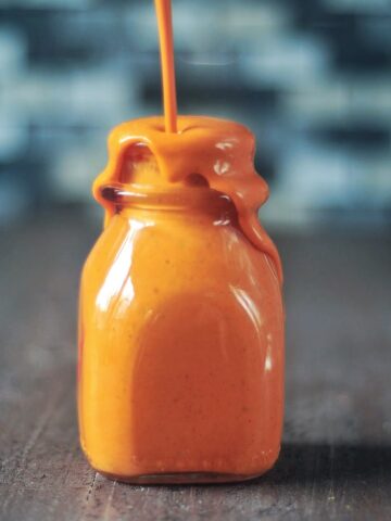 A small glass bottle with bright orange hot sauce being poured in until it is overflowing out the top. Bottle sits on a wood surface with a tile backsplash blurred in background.