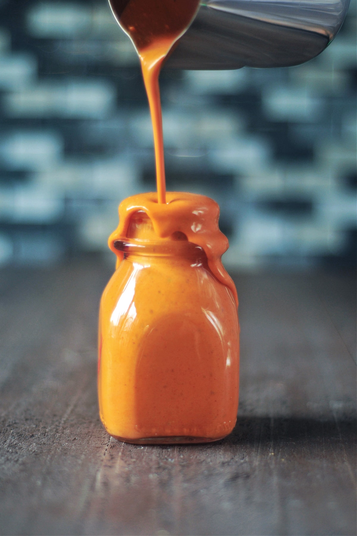 A small glass bottle with bright orange hot sauce being poured in until it is overflowing out the top. Bottle sits on a wood surface with a tile backsplash blurred in background.