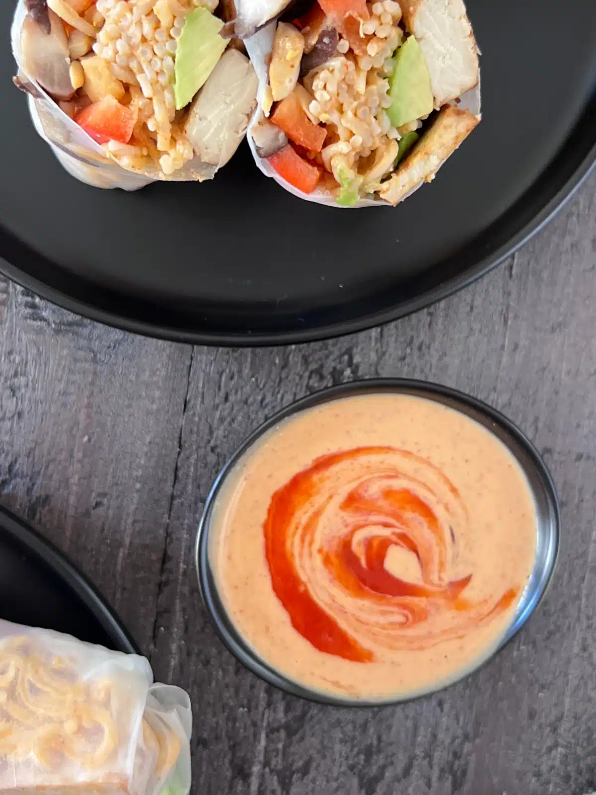 A small bowl of spicy almond butter sauce sits next to a plate of avocado shiitake spring rolls.